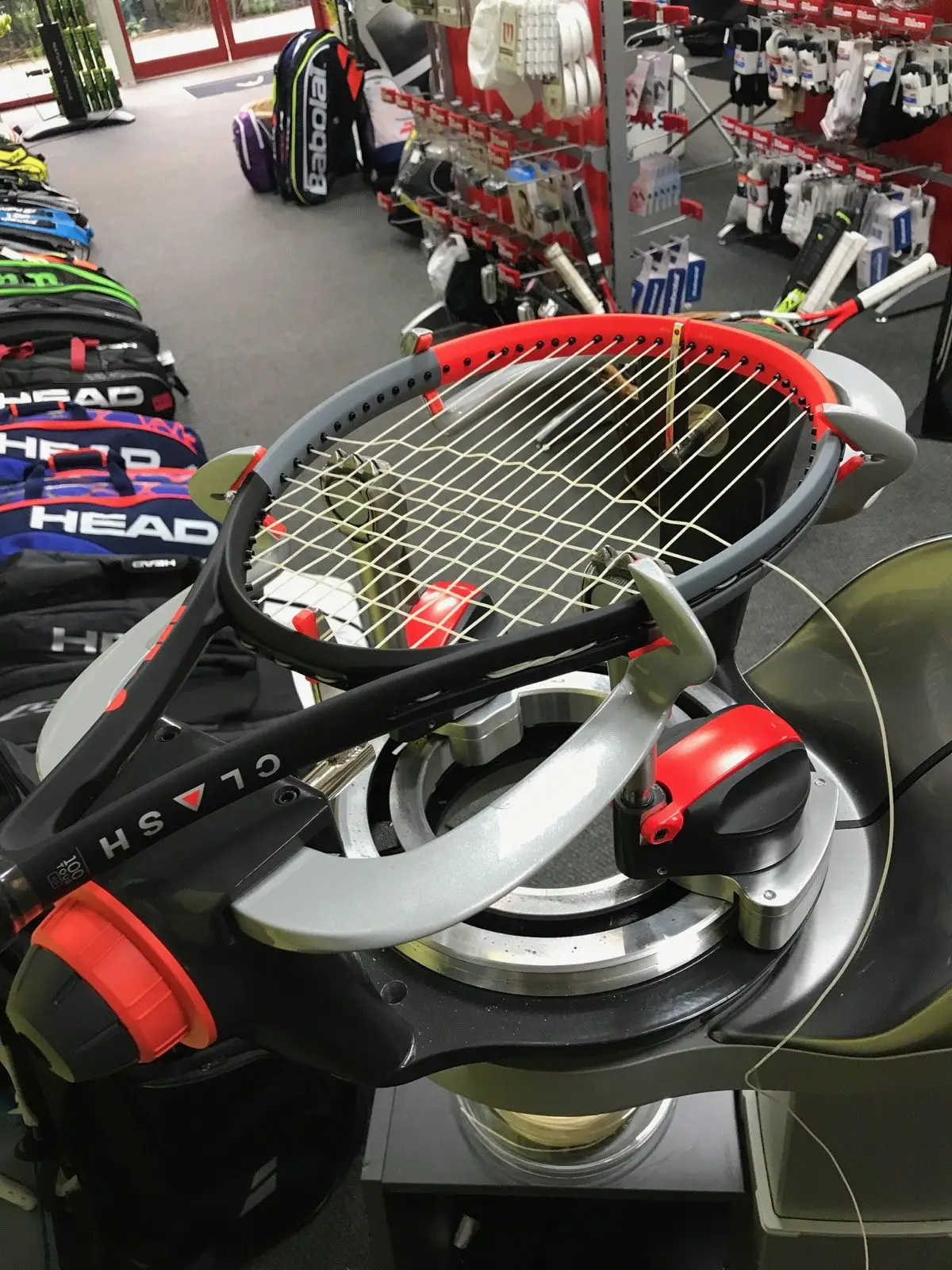 tennis racquet being restrung on a restringing machine in a shop with other tennis and racquet sports equipment in the background